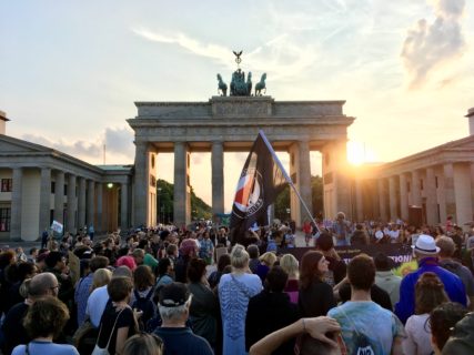 A crowd stands in front of Brandenburg Gate with the sun setting behind the gate