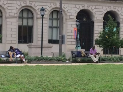 Two men sit on two different benches in front of the library. One is sleeping. He leans on bags that are full and a cane is outstreched beside his legs. The other sits up, with his hands clasped between his legs and head turned down. A backpack and several other bags sit next to him on the bench.