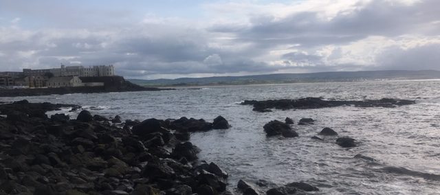 A rocky beach in the foreground on a cloudy day, with buildings in the background on an outcrop.