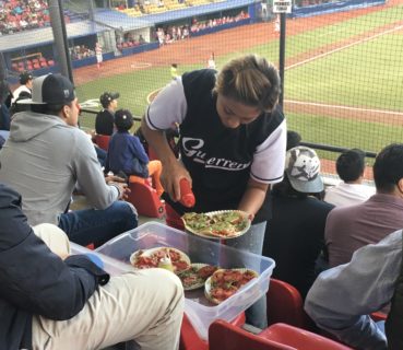 A female concession vendor wearing a Guerreros baseball jersey sells tostadas to fans.