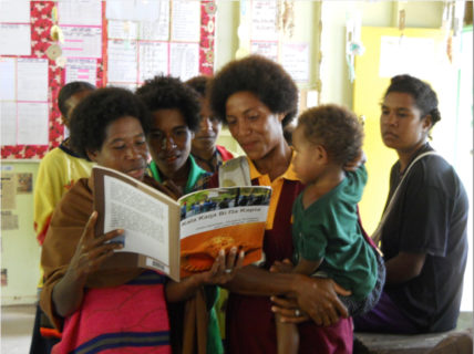 A photo of one woman holding open a book while two others look on, one of whom is holding a young child in her arms.