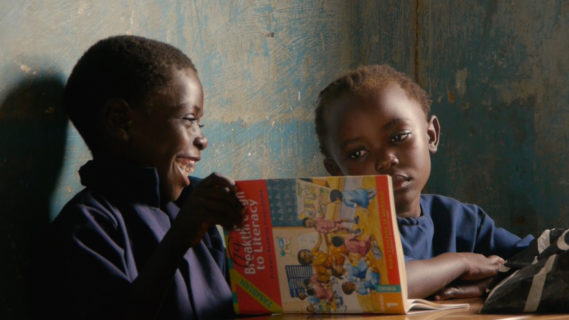 A photo of two children sitting next to each other, the boy smiles with an open book in his hands, while the girl looks over at him.