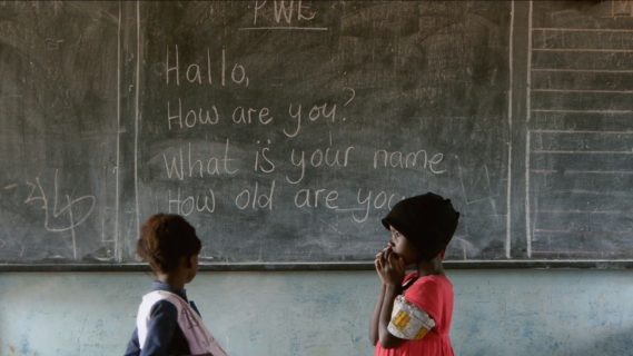 Two children stand across from each other in front of a chalkboard with writing on it that reads "Hallo, How are you? What is your name How old are you."