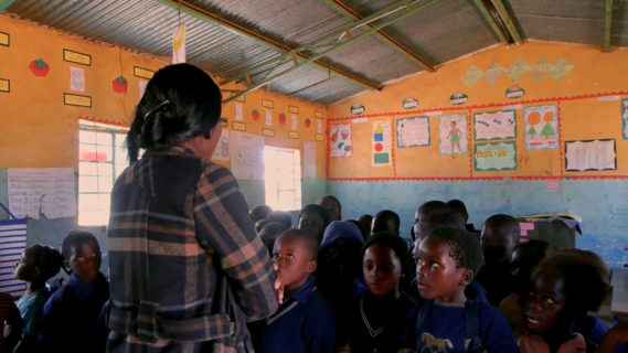 A photo of children facing the camera in a classroom with their teacher standing in front of them.