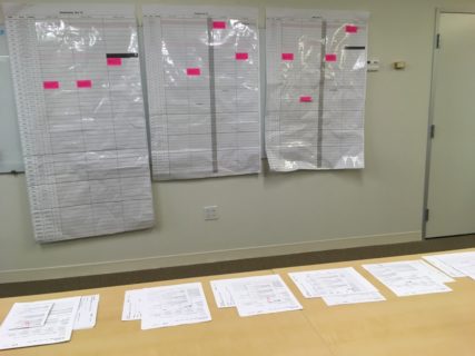 Five piles of paper lie on a table top in the foreground. Behind these three large pieces of paper hang on a wall. They are covered with a grid and marked with a few pink rectangular stickers.