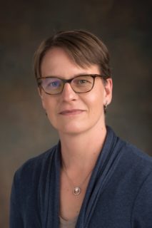 A woman with short brown hair looks straight ahead. She is wearing a gold necklace with pendant and a v-neck navy blouse