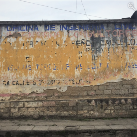 A damaged adobe wall that is completely covered with a faded sign with blue and black words and yellow and white background.