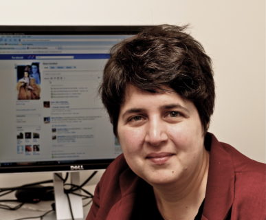 A photo of an individual with short brown hair smiling at the camera and sitting in front of a computer screen.