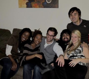 A photo of six people sitting on a couch leaning against each other and smiling.