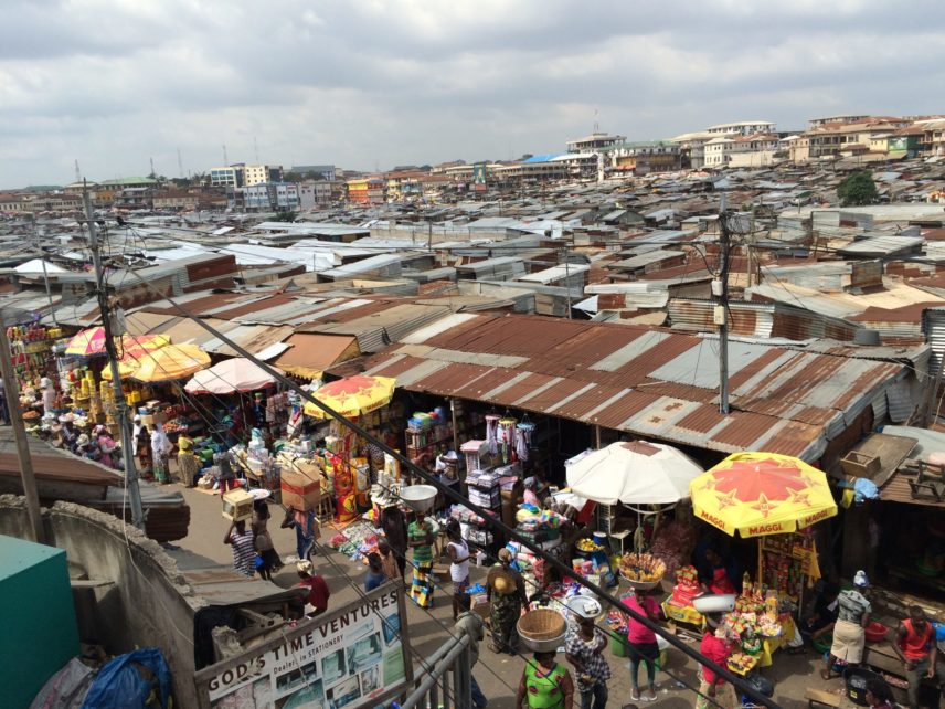 Men and women carry baskets and boxes of goods on their heads on a busy street in Kumasi Central Market.