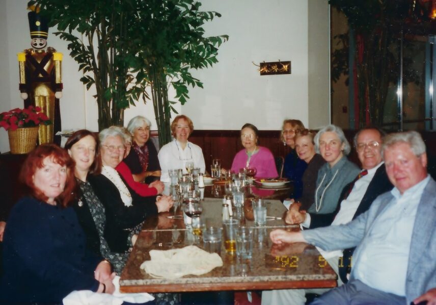 Photograph of a number of men and women, grouped around a table laden with glasses.