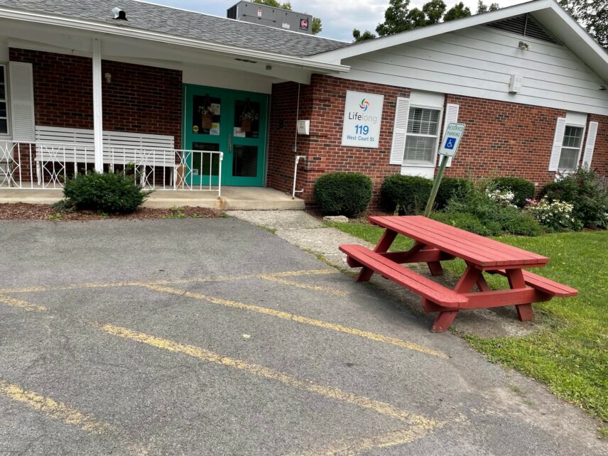 Photograph of exterior of brick building with green door and picnic table in foreground.