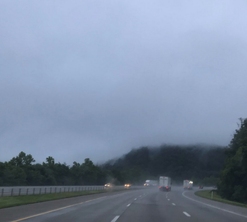 Photograph of a highway with cars and semi-trucks on it, gray skies and green hills in the distance.