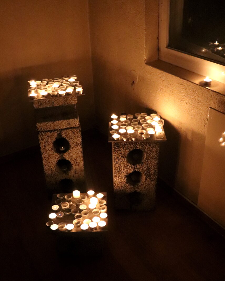 Photograph of votive candles, some lit and others burned out, positioned on three cement blocks of various sizes.