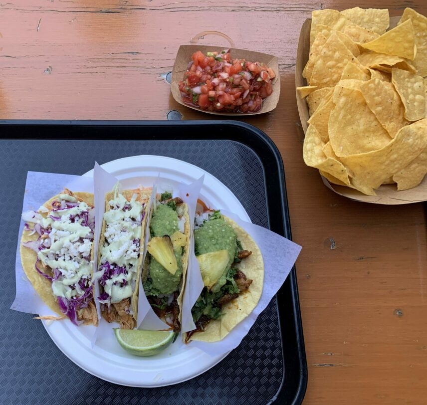 Four tacos on a plate on a wooden table with a lime wedge; two are al pastor pork with pineapple slices and a salsa verde, and two are slow-cooked chicken topped with purple cabbage and a serrano-cream sauce. Also on the table are golden-yellow corn tortilla chips and a pico de gallo of tomatoes, red onion, lime juice and jalapeños.