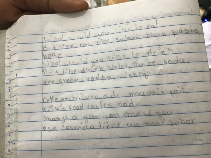 A student’s hand appears in the left corner, holding a notebook with white-lined pages. In pencil, the student has written a question-and-answer series in English to ask what a customer would like to eat and drink. The student has also written “the food tastes bad” in French, English, Kreyòl, and Spanish.