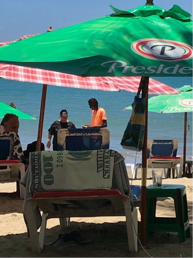 A tall Dominican male worker with shoulder-length braids and a bright orange t-shirt holds a paper and pen while talking to a shorter female tourist wearing a black beach dress, glasses, and a ponytail. They are standing on the sand in front of the ocean. They are behind three blue lawn chairs covered by green beach umbrellas with the Presidente beer logo. An empty lawn chair is covered by a towel printed with a one-hundred-dollar bill.