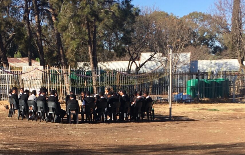 A group of students and their teacher sit on black plastic chairs in a circle on a dirt field. They are participating in a discussion. Their school building can be seen in the background as well as several green water storage tanks.