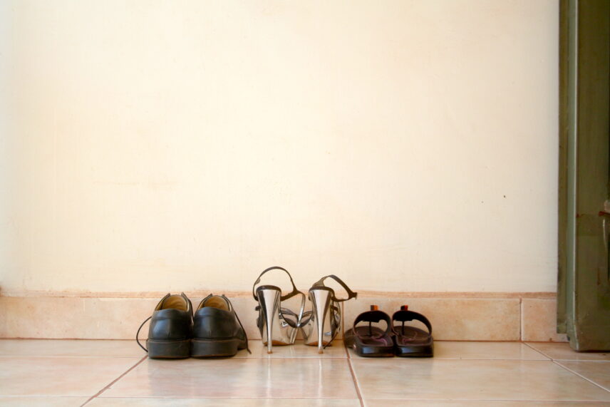 Three pairs of shoes are lined up against a wall on a tiled floor. The first is a black pair of shoes, the second is a pair of shiny silver high heeled sandals, and the third is a pair of black sandals.