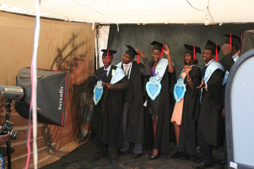 In front of a professional camera, a group of six students, both men and women, stand together in graduation caps and gowns, preparing for a photograph.