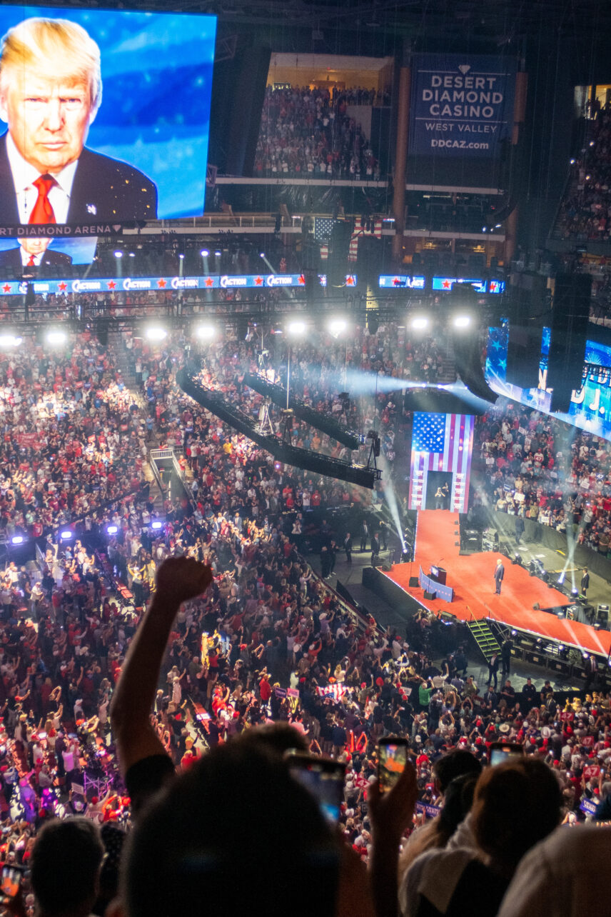 Image of the inside of a large stadium full of people with a stage observable in the lower right which features Former President Donald J. Trump. In the upper left corner there is an image of a large screen in the stadium showing an image of Mr. Trump.