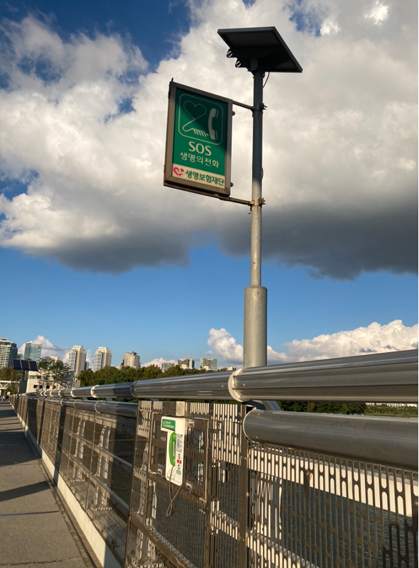 Alt text: An SOS hotline phone on a bridge over a river with a view of city buildings in the background, unrelated to Yuna’s crisis experience.