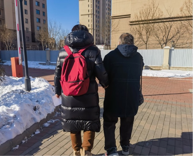 A photograph showing a young woman holding hand with an older woman. There are piles of snow at the sides of the walkway.
