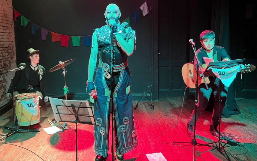Picture showing the three members of Somos Mink’a Folklore Queer group on stage, Dany Vila playing the drum, Legon Queen singing, and Macu playing the guitar
