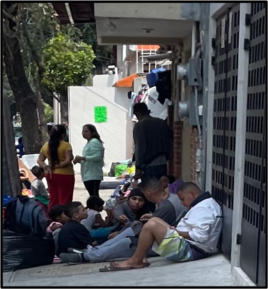 A group of several migrants, adults and children. are sitting on the ground looking at a telephone. Two women are standing behind them, having a conversation.