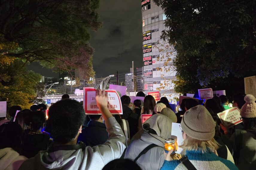A large group of protesters with their backs to the viewer carrying small placards. Tall buildings are visible in the distance.