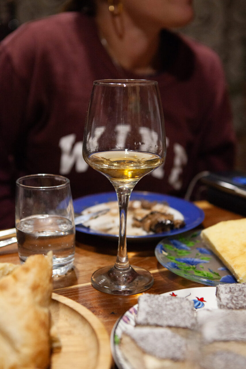 A glass of wine sits on a table with food. In the background is a person wearing a sweatshirt that reads, “WINE.”