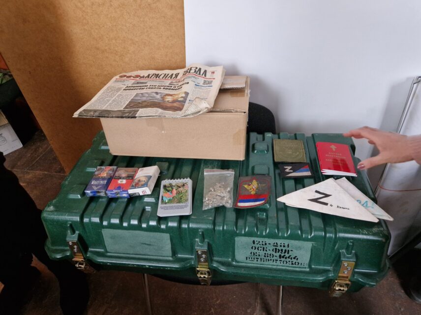 A green suitcase lies in the centre of the image, three packs of cigarettes are played on top of it on the left, along with Russian military armbands, pins and prayer books. Many items display a ‘Z’, the symbol of pro-war sentiment in Russia.