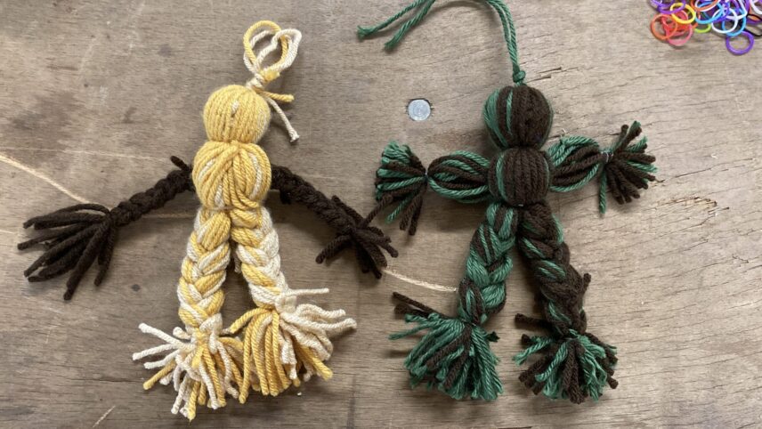 Two dolls made of fabric. The doll on the left has a two-toned yellow body. Its arms are made brown braids, while its legs are made of yellow braids. The doll on the right is similar in composition but made of green and brown braids.