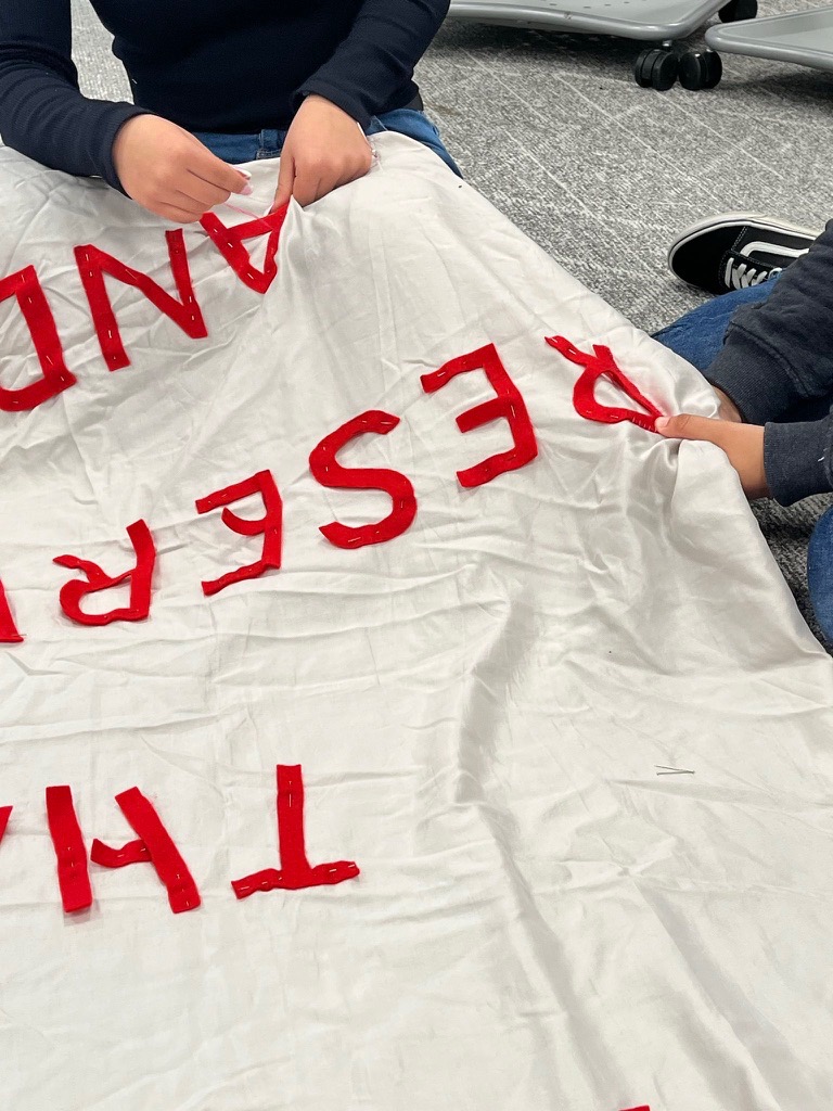 Two students hands can be seen sowing red fabric letters.