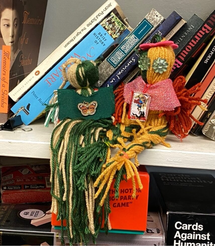 Two dolls sit side by side on a bookshelf. The doll on the left is made of light brown and green fabric and wears a green make-shift shirt with a butterfly charm. The doll on the right has jade-colored flower eyes and is made of yellow and green fabric. It wears a pink glitter shirt and a El Niño necklace.