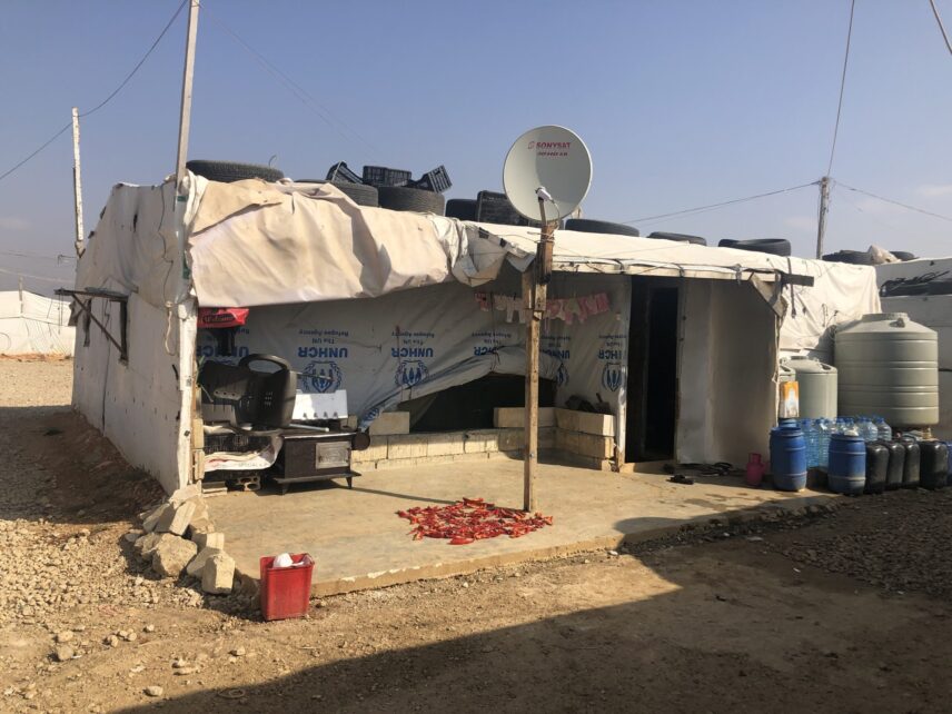A tent constructed with UNHCR-branded tarp and cinder blocks. A satellite dish labeled is mounted on a wooden pole at the front. Red peppers are laid out to dry on the concrete patio and bottles of cooking gas are stacked along the right side near large water tanks. Laundry is hanging on a line under the awning, and the ground around the tent is covered in gravel.