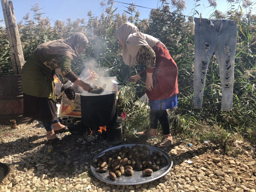 Two women wearing headscarves cook over an open fire outdoors. Both are learned over a large, steaming black pot. A metal tray filled with eggplants sits on the ground in front of them. They are surrounded by a tall thicket of grass, and to the right, a pair of ripped jeans hangs on a clothesline.