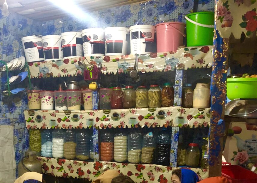 A vibrant and densely packed kitchen pantry shelf covered in floral and coffee-themed plastic linings. The shelves are filled with a variety of plastic and glass jars containing pickled vegetables, spices, grains, and legumes.