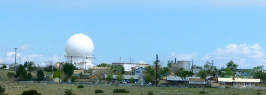 A large white radar dome rises above a small community with modest houses and trees, under a bright blue sky.