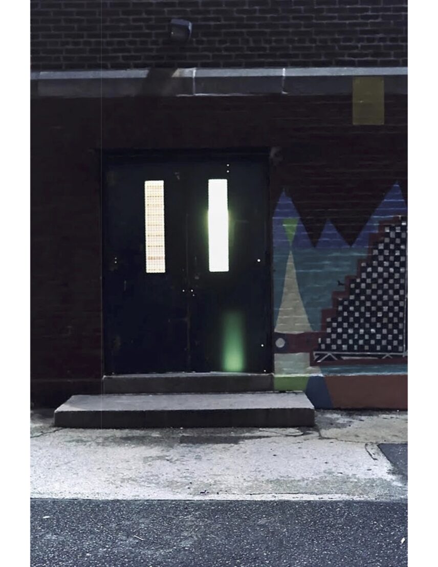 A nighttime photo of two school doors with small rectangular windows and a wall with colorful graffiti. There is florescent lighting from a stairwell behind the school doors.