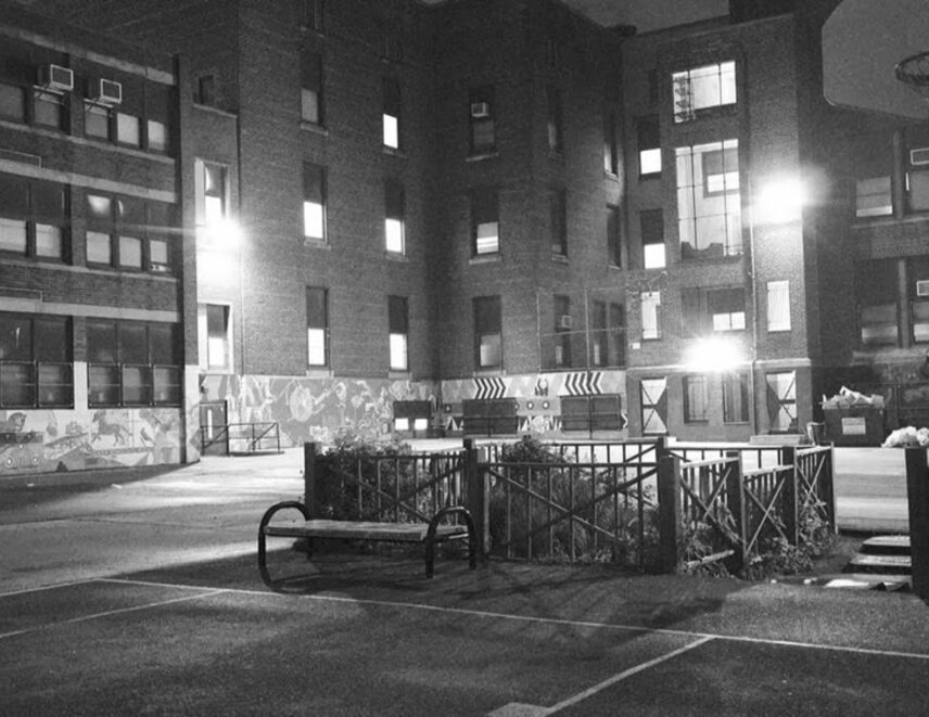 A black and white photo of a playground with a school in the background. On the right side, there is a small garden, a basketball hoop, and a bench.
