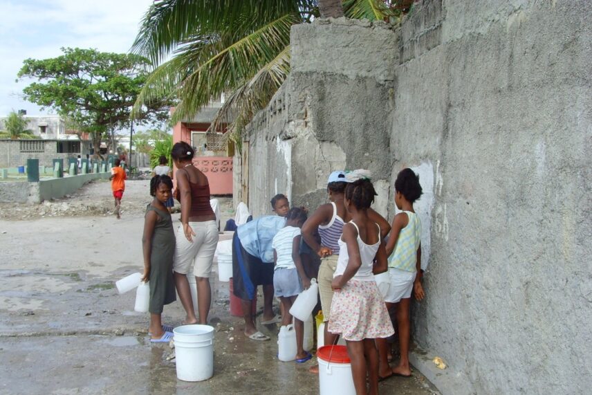 Eight Haitian children of varied ages are standing outside on wet ground near a wall. They are gathered around a water source with large white buckets and other receptacles.
