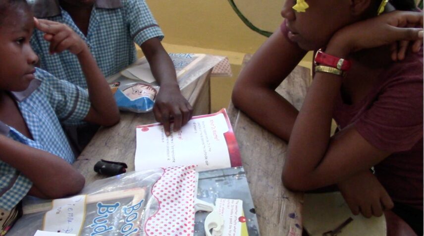 Three children are sitting around a table with a school book and other school supplies in the middle. One child has a finger placed on the book, pointing to a word. Another child has two fingers placed on her forehead.