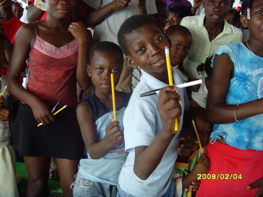 A group of Haitian children of different ages fills the frame. The boy in the front of the group holds a cross made from a vertical pencil and a horizontal ink pen. A smaller boy behind him holds up a pencil.