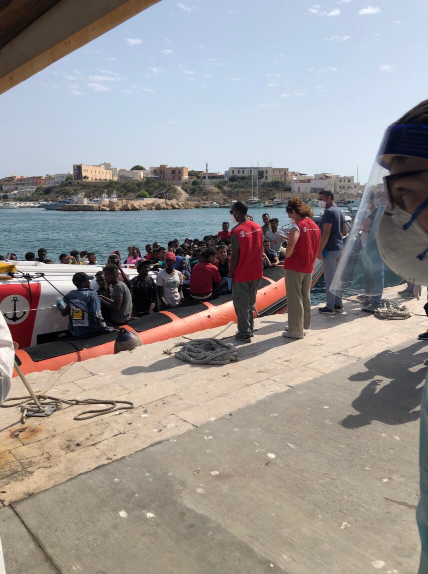 A boat, with migrants sitting in it, arrives at the port. Two people wearing red vests receive the boat.