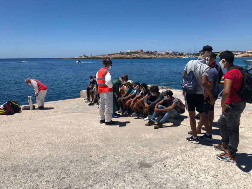 A queue of migrants squat the port at the edge of the water. A doctor in a white shirt, khaki pants, and a red vest evaluates the migrants.