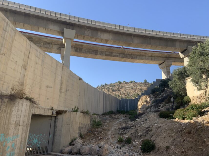 photo of the wall, concrete and some ten meters high, cutting through Bir Ona with a closed metal gate covered in teal handprints and other graffiti. A two-lane highway bisects the skyline above. The edge of a building and some shrubbery enters the righthand side of the frame. The olive orchards are visible on a hill behind the wall.