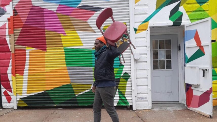a man wearing black clothing and an orange cap carrying a red chair over his shoulder, walks past a colorful mural and an open white door, appearing to be in motion.