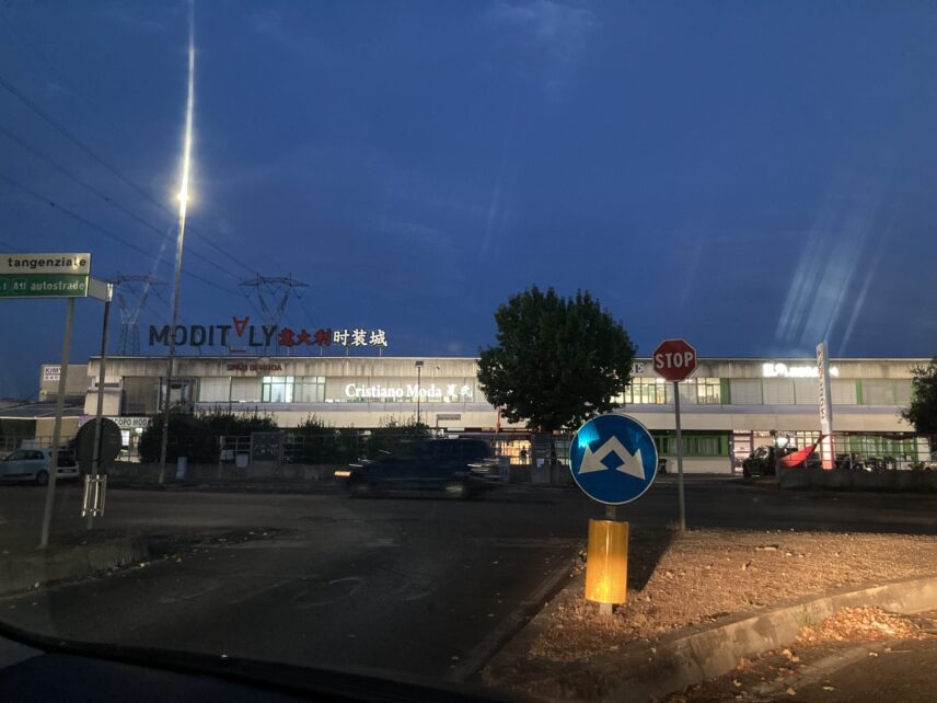 Suburban commercial building in Prato with illuminated signs in Italian and Chinese advertising fashion businesses.