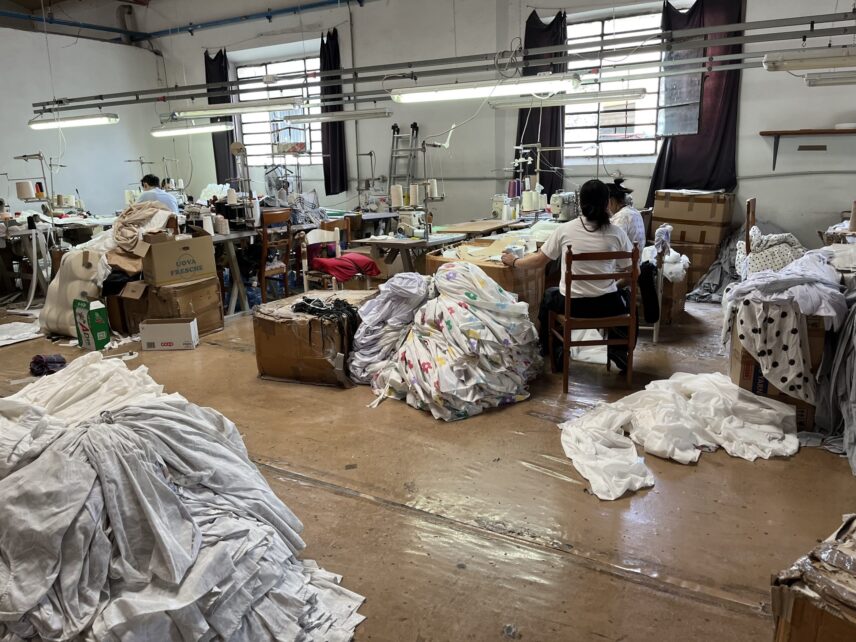 Interior of a sewing workshop with workers at machines and piles of garments on the floor.
